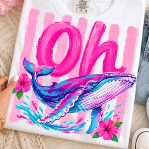 Pu&ograve; includere: Una t-shirt bianca con una grande scritta rosa a pennellata che recita Oh, accompagnata da una balena blu e viola. Il design presenta strisce verticali rosa, fiori di ibisco rosa acceso e spruzzi d'acqua blu sulla parte anteriore.