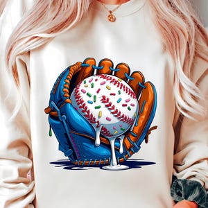 May include: A cream-coloured sweatshirt featuring a baseball glove in blue and brown, holding a baseball covered in colourful sprinkles and dripping white icing. The baseball has red stitching. The design is a playful take on a classic sport.