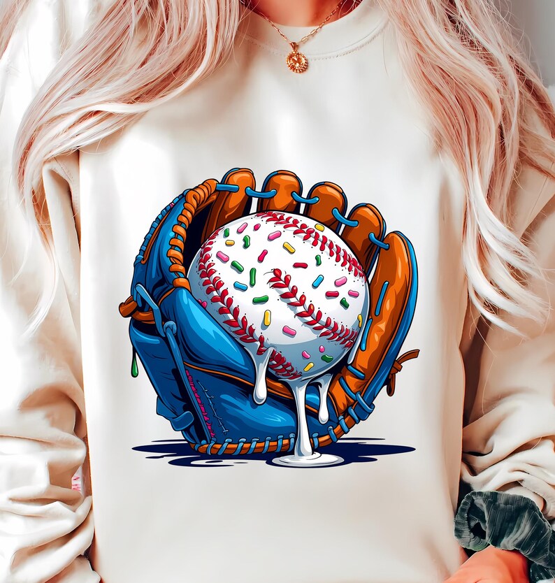 May include: A cream-coloured sweatshirt featuring a baseball glove in blue and brown, holding a baseball covered in colourful sprinkles and dripping white icing. The baseball has red stitching. The design is a playful take on a classic sport.
