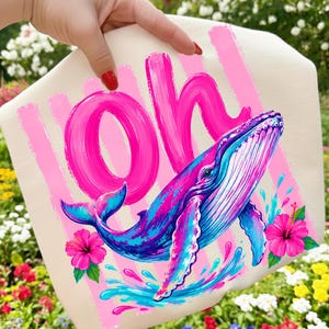Pu&ograve; includere: Una borsa tote color crema con una grafica vivace di una megattera blu e viola che salta fuori dall'acqua. Il design include la parola "Oh" in grandi lettere rosa brillante effetto pennellata, accentuata da due fiori di ibisco rosa.