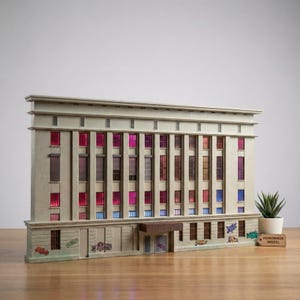 Könnte beinhalten: Ein handgefertigtes Modell eines Gebäudes mit mehreren Fenstern, einige mit farbenfrohen rosa, roten und blauen Akzenten. Das Gebäude hat eine hellbeige Fassade mit Graffiti-Kunst auf der unteren Ebene. Eine kleine Topfpflanze und ein Etikett mit der Aufschrift "HANDMADE MODEL" befinden sich daneben.