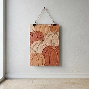 May include: A rectangular wall hanging featuring a textured impasto painting of multiple pumpkins in shades of cream, beige, tan, and burnt orange. The artwork is suspended by a thin rope attached to two black metal clips at the top edge.