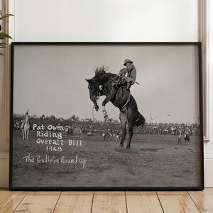 Puede incluir: Una impresión enmarcada en blanco y negro que representa una escena de rodeo de 1928. La imagen muestra a un jinete sobre un caballo encabritado con el texto: Pat Owens Riding Overall Bill 1928 The Pendleton Round up. La impresión tiene un marco negro.