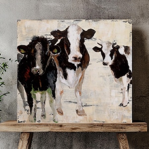 Puede incluir: Cuadro cuadrado que representa tres vacas en un estilo rústico. Las vacas están representadas en blanco y negro con detalles en marrón, sobre un fondo texturizado blanquecino. La obra de arte se exhibe sobre un banco de madera, con una planta en maceta en primer plano.