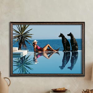 Pode incluir: Uma mulher de biquíni rosa e chapéu branco relaxa à beira de uma piscina com dois cães pretos. A piscina é cercada por um deck de azulejos brancos e uma palmeira. A água é azul e reflete o céu e a mulher.