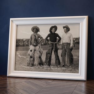 Puede incluir: Una fotografía en blanco y negro enmarcada que muestra a tres personas con vestimenta del oeste de pie en un campo. El texto dice: Sunhouse, Hori, Riffle, Lee Moorhouse, Champions of the World, Pendleton, Ore, 1916. El marco es blanco con molduras.