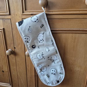 May include: A beige double oven glove featuring a repeating pattern of white sheep with black details. The glove is hanging on a wooden cabinet. The sheep design includes various poses and expressions.