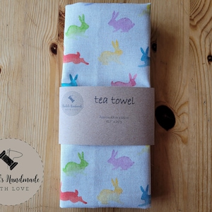May include: A folded tea towel with a light blue background and a pattern of colorful bunny rabbits. The towel is wrapped with a brown paper band that reads "tea towel". The towel measures approximately 43cm x 66cm.