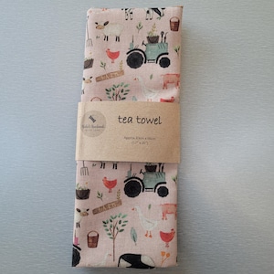 May include: A pink tea towel with a farm theme. The towel features a tractor, chickens, pigs, sheep, and other farm animals. The text "tea towel" and "Approx 41cm x 66cm (16" x 26")" is printed on the label.