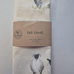May include: A folded tea towel featuring a sheep design. The beige towel has a brown paper band that reads "tea towel" and "Hebels Handmade with Love." The towel measures approximately 43cm x 66cm.