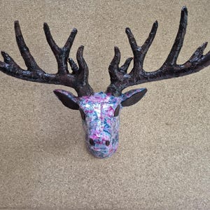 May include: A decorative deer head wall mount with large, dark brown antlers. The deer's head is patterned with a floral design in shades of pink, blue, and white. The ears are dark brown, creating a rustic, artistic wall decoration.