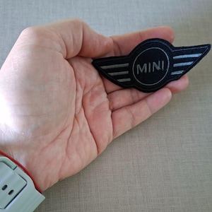 Puede incluir: Un parche con el logotipo MINI negro y plateado con la palabra "MINI" en un círculo blanco. El parche tiene un diseño de ala a cada lado. El parche se sostiene en una mano.