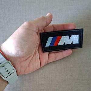 Puede incluir: Parche rectangular negro con las letras "M" y un diseño de rayas azules, rojas y celeste. El parche se sostiene en una mano, con un reloj gris en la muñeca.