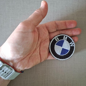 Puede incluir: Una taza de cerámica blanca con un asa negra y el texto "But First, Coffee" en negrita negra. La taza está sobre una superficie de madera. La taza mide aproximadamente 10 cm de altura.