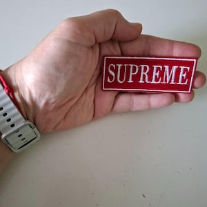 May include: A rectangular red patch with the word "SUPREME" embroidered in white capital letters. The patch is held in a hand, and the background is a light-colored surface.