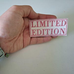 Könnte beinhalten: Eine weiße Uhr mit hellgrauem Armband wird am Handgelenk getragen. Ein rechteckiger Aufnäher mit den Worten "LIMITED EDITION" in rosa Schrift wird in einer Hand gehalten.