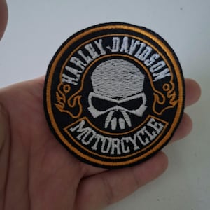 Puede incluir: Parche bordado redondo con un diseño de calavera y las palabras "HARLEY-DAVIDSON MOTORCYCLE". El parche tiene un fondo negro con detalles blancos y amarillos. El parche es circular.