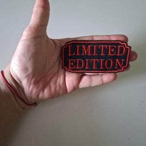 Könnte beinhalten: Ein schwarzer rechteckiger Aufnäher mit rotem Rand und den Worten "LIMITED EDITION" in Rot gestickt. Der Aufnäher wird in einer Hand gehalten, um seine Größe und sein Design zu zeigen. Der Aufnäher ist wahrscheinlich für Kleidung oder Accessoires.