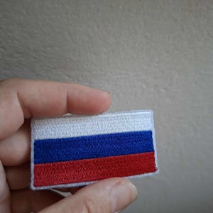 May include: A rectangular patch depicting the Russian flag. The design includes three horizontal stripes: white above, blue in the centre, and red below. The patch is probably made of fabric and has a stitched border.