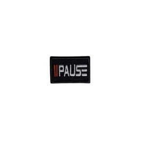 May include: A rectangular black patch with the word "PAUSE" embroidered in white. The word is flanked by two vertical red stripes on the left and two horizontal white stripes on the right. The patch is likely for clothing or accessories.