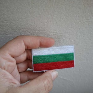 Puede incluir: Un parche rectangular con la bandera de Bulgaria. La bandera consta de tres franjas horizontales: blanca, verde y roja. El parche, probablemente de tela, se muestra sobre un fondo neutro.