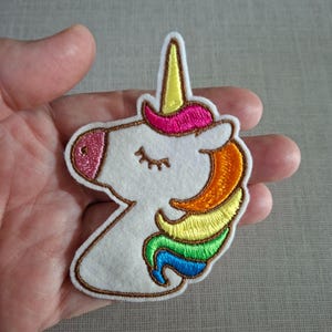 Könnte beinhalten: Ein weißer Einhorn-Flicken aus Filz mit einer Regenbogenmähne und einem goldenen Horn. Das Einhorn hat eine rosa Glitzer-Nase und geschlossene Augen. Der Flicken ist braun umrandet und hat einen weißen Rand.