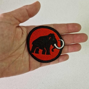 Op de afbeelding: Geborduurde patch met een zwart silhouet van een mammoet op een rode cirkelvormige achtergrond. De mammoet heeft een witte slagtand en een zwarte rand. Geschikt voor jassen, tassen of andere textielartikelen.