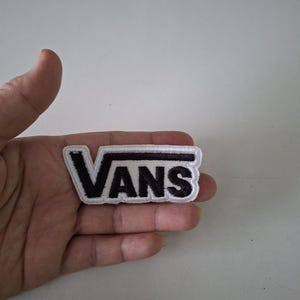 May include: A black and white Vans logo patch. The patch features the word "VANS" in a bold, sans-serif font, with a white border. The patch is rectangular and appears to be made of fabric.