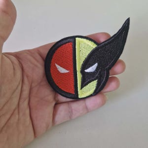 Peut inclure: Écusson brodé avec un design divisé : une moitié rouge avec un œil blanc, l'autre moitié jaune avec un masque noir et un œil blanc. L'écusson a une bordure noire, pour vêtements ou accessoires.