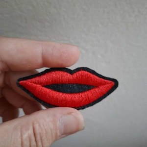 May include: Embroidered patch featuring a pair of red lips with a black outline. The lips are a vibrant red color, and the patch has a black border. This patch is suitable for clothing or accessories.