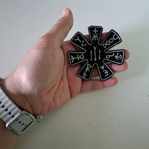 Puede incluir: Parche negro en forma de estrella con símbolos bordados en blanco y borde blanco. El parche, sostenido en una mano, tiene un diseño central de líneas verticales. Probablemente sea un emblema o un elemento decorativo.