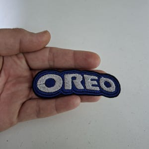 May include: Embroidered patch with the word "OREO" in white stitching on a blue and black background. The patch is oval and has a stitched border. The patch is held in a hand.