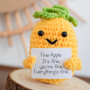 Op de afbeelding: Een gehaakte gele ananasfiguur met een groene bladtop en zwarte kraaloogjes. De figuur houdt een wit bord vast met de tekst "Fine-Apple, It's fine, you're fine, Everything's fine."
