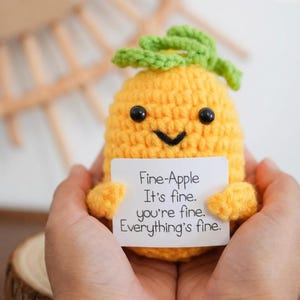 May include: A crocheted yellow pineapple with a green leafy top and black eyes. The pineapple holds a white card with the text "Fine-Apple It's fine. you're fine. Everything's fine."