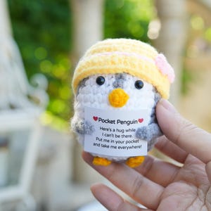 Könnte beinhalten: Ein gehäkeltes Pinguin-Spielzeug mit gelbem Hut und rosa Blume. Der Pinguin hat einen weißen Körper, graue Flügel und einen orangefarbenen Schnabel und Füße. Er hält ein Schild mit der Aufschrift "Pocket Penguin".
