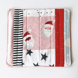 May include: A pink, white, red, and blue quilted pot holder with a patchwork design featuring Santa Claus faces and stars.