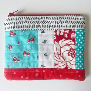 May include: A quilted pouch with a red zipper and a variety of fabric patterns. The pouch features sections of turquoise with mushroom designs, white with black stars, red floral, and turquoise polka dots. The top and bottom edges are red.