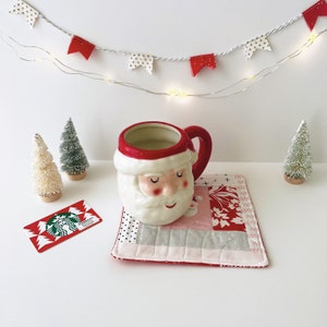 May include: A white ceramic Santa Claus mug with a red hat and a pink face, sitting on a pink and red patchwork coaster. A Starbucks gift card is on the table next to the mug.