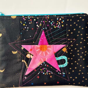 Peut inclure: Pochette zippée noire avec un grand motif d'étoile rose. L'étoile est superposée et cousue sur le tissu noir, décoré d'étoiles dorées et de motifs célestes. La fermeture éclair est turquoise.