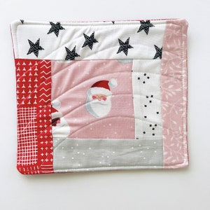 May include: A pink and red quilted pot holder with a Santa Claus design and a white background. The pot holder is made of fabric and has a quilted design.