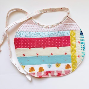 May include: A handmade, quilted baby bib with a variety of colorful fabric patterns. The bib features stripes of pink hearts, blue polka dots, red dots, and floral designs. The bib has a rounded shape and white ties with small pink dots.