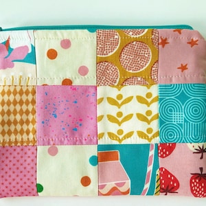 Puede incluir: Un estuche rectangular con cremallera confeccionado con un patchwork de diversas telas de algodón. El diseño presenta una cuadrícula de cuadrados con patrones variados que incluyen fresas rosas, círculos geométricos turquesa y lunares.