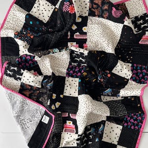 May include: A black and white patchwork quilt with a pink binding. The quilt is made of various fabrics with different patterns, including stars, polka dots, and geometric shapes.