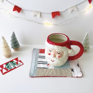 以下が含まれることがあります： 白いひげと目を閉じた赤いサンタクロースのマグカップが、パッチワークのコースターの上に置かれています。マグカップには白い液体が注がれています。赤い旗と金の旗がついた白い電飾がマグカップの上にぶら下がっています。