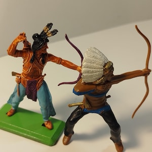 May include: Two toy Native American figures in a battle scene. One figure is in orange and blue clothing, with a black headdress and a raised arm. The other figure is aiming a bow and arrow, wearing a white feathered headdress and black trousers.