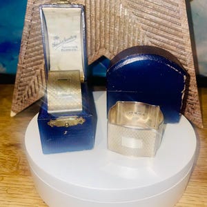 Könnte beinhalten: Zwei silberne Serviettenringe, einer in einer blauen Schmuckschatulle. Die Schatulle ist geöffnet und zeigt einen goldfarbenen Innenraum. Der andere Serviettenring befindet sich außerhalb der Schatulle. Die Schatullen tragen den Text "The Fabulous Jewellers, Burnley".