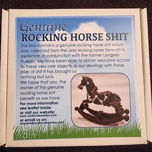 Puede incluir: Una caja blanca con el texto "Genuine ROCKING HORSE SHIT" en azul. La caja contiene un dibujo de un caballo balancín marrón y blanco. El texto también incluye una dirección de sitio web.