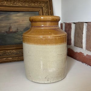 May include: A cylindrical stoneware crock featuring a two-tone glaze with a glossy brown upper section and a matte cream-coloured lower body. The vessel has a thick, rounded rim and a flat base, displaying subtle surface textures and markings.