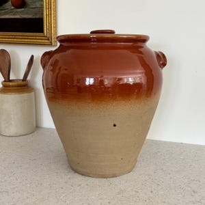 May include: A large, glazed ceramic jar with a lid. The jar is a rich brown colour at the top, fading to a tan colour at the base. It has two small handles on the sides and a small knob on the lid. The jar is likely for storage.