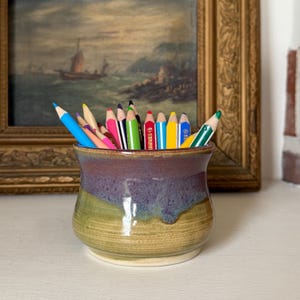 May include: A ceramic pencil holder with a flared rim and a two-tone glaze featuring a deep purple top section that transitions into a textured olive green base. The holder is filled with a collection of sharpened coloured pencils in various hues.
